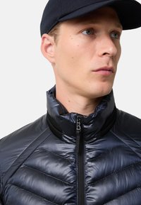 Námořnický modrý péřový kabát s vysokým límcem, který má horizontálně prošívanou texturu a přední zip. Síťové akcenty v oblasti krku.