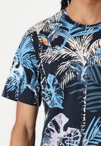 Navy kortermet T-skjorte med et tropisk bladmønster i lys blått og rosa, med elementer av fargerike fugler. Stoffet ser mykt ut.