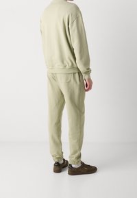 Man draagt lichtbeige sweatshirt en joggers met elastische boorden en donkerbruine sneakers, staand tegen een effen witte achtergrond.
