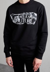 Svart sweatshirt av bomullsblandning med en sliten vit grafisk tryck med texten "Versace Jeans Couture". Ribbstickade muddar och fåll.