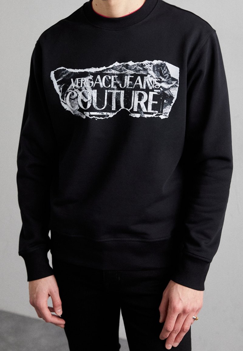Svart sweatshirt av bomullsblandning med en sliten vit grafisk tryck med texten "Versace Jeans Couture". Ribbstickade muddar och fåll.