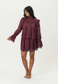 Robe en dentelle bordeaux avec un design à volants, des manches ondulées et un col haut. La texture transparente et l'ourlet festonné accentuent la silhouette.
