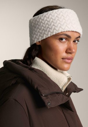 Bandeau en tricot blanc avec un motif en chevrons, porté avec une veste matelassée marron et un pull crème à col montant. Texture lisse.