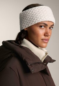 Bandeau en tricot blanc avec un motif en chevrons, porté avec une veste matelassée marron et un pull crème à col montant. Texture lisse.