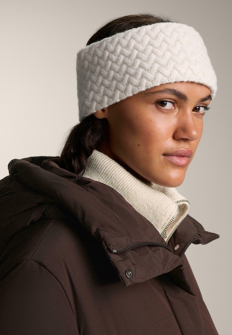 Bandeau en tricot blanc avec un motif en chevrons, porté avec une veste matelassée marron et un pull crème à col montant. Texture lisse.