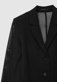 Blazer nero con colletto strutturato, due bottoni neri e maniche trasparenti. Il tessuto ha una texture liscia con una vestibilità sartoriale.