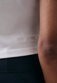 Fehér Dri-FIT póló, sima textúrával, emblémázott márkával az alján. Fekete sportleggings félig látható.