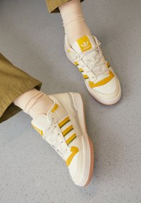 Zapatillas blancas con rayas amarillas y verdes, que presentan un diseño texturizado, material de cuero suave, suela de goma y acentos perforados.