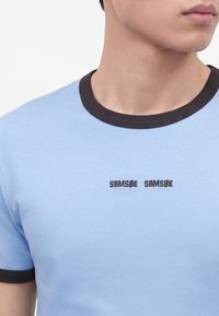 Homme portant un t-shirt bleu clair avec col et bords de manches noirs, avec le texte « "SAMSØE SAMSØE" » sur la poitrine.
