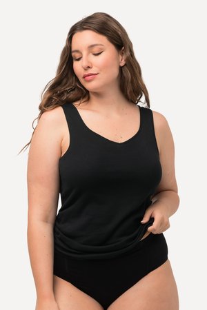 Donna con lunghi capelli castani che indossa un top nero senza maniche e biancheria intima nera, tirando delicatamente l'orlo del top mentre guarda verso il basso.