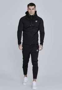Czarna hoodie z zamkiem na całej długości, z regulowanymi sznureczkami oraz dopasowane spodnie dresowe z logotypem. Całość ma dopasowany krój i wykonana jest z gładkiego, rozciągliwego materiału.