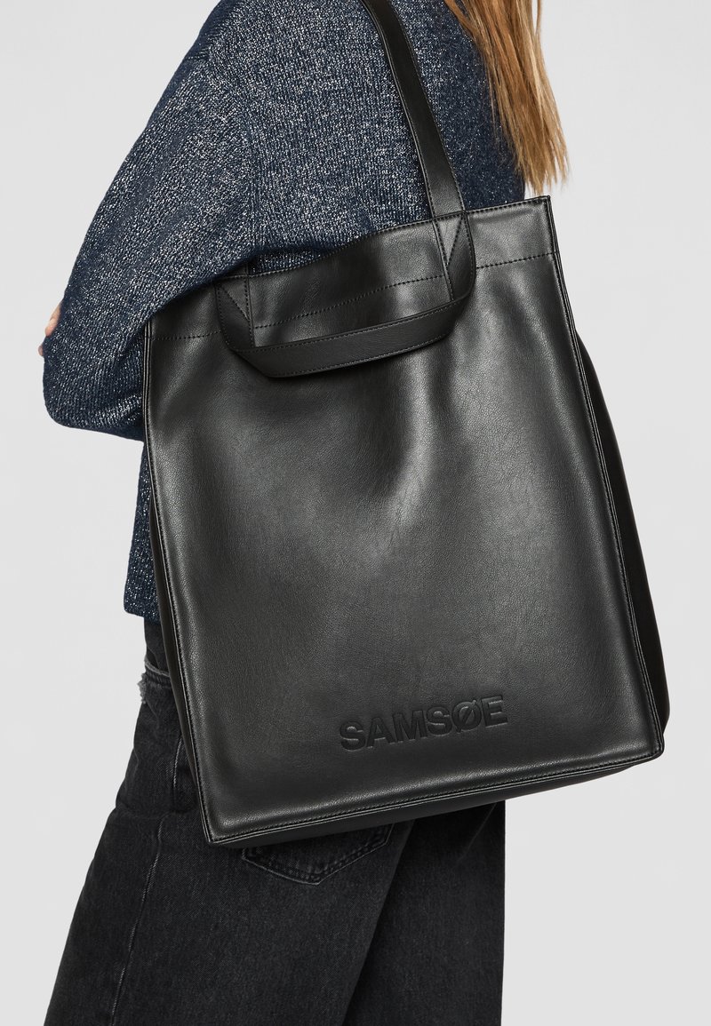 Sac fourre-tout en cuir noir au design simple, avec une anse plate et un logo embossé. Texture lisse avec une forme structurée.