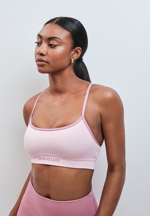 Femme aux cheveux foncés attachés, portant un brassière de sport rose clair et un legging taille haute, debout contre un mur clair uni.