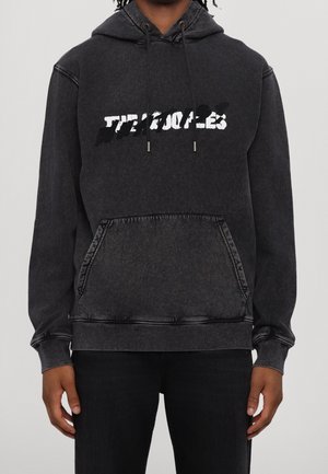 Osoba nosząca wypraną czarną bluzę z kapturem z kieszenią kangurka z przodu oraz częściowo zasłonięty biało-czarny napis na klatce piersiowej.