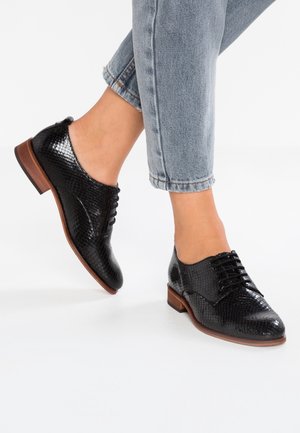 Czarne buty skórzane w wzór wężowej skóry z niskim obcasem, z sznurówkami i zaokrąglonym noskiem, noszone z dżinsami o długości kostki.