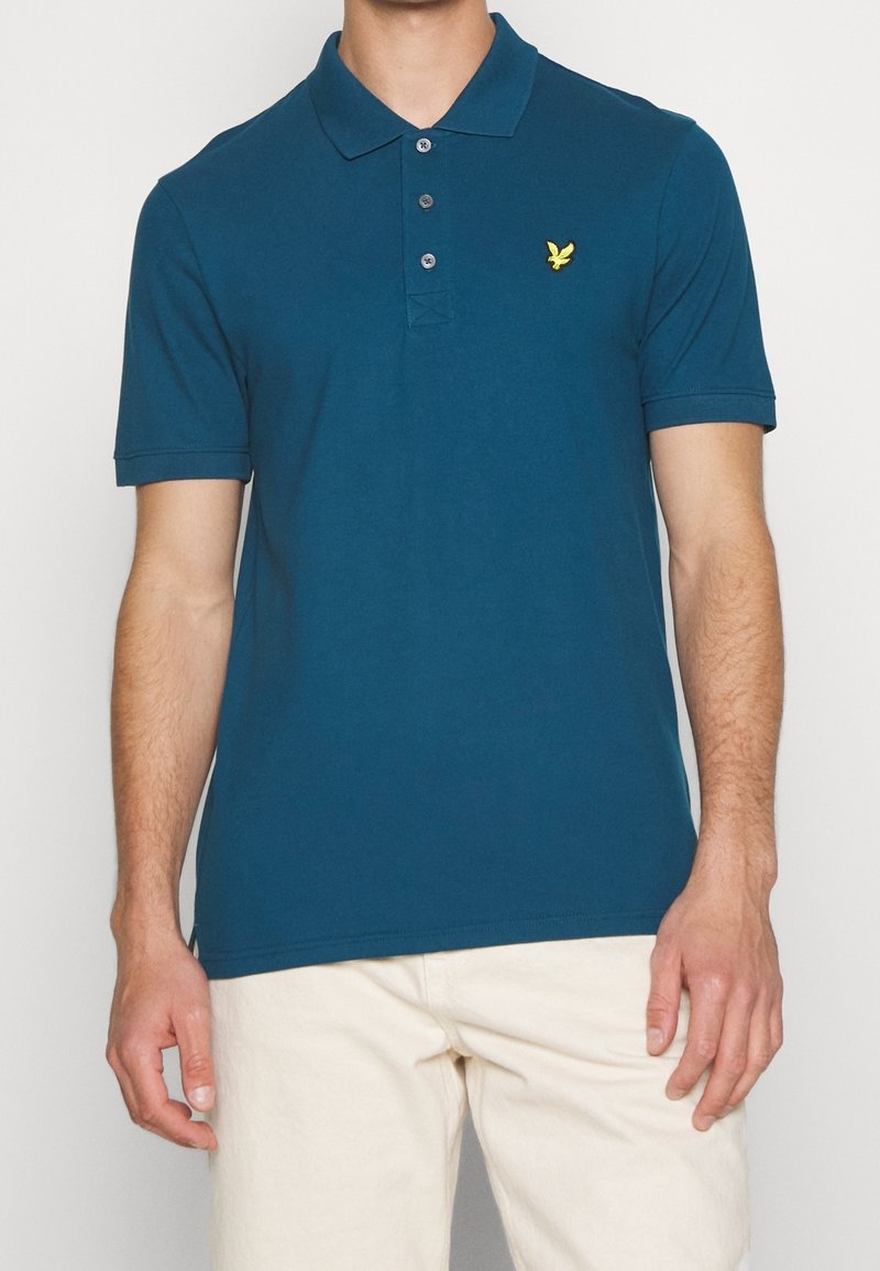Homme portant un polo bleu sarcelle à manches courtes avec trois boutons et un petit logo d'oiseau jaune sur la poitrine gauche.