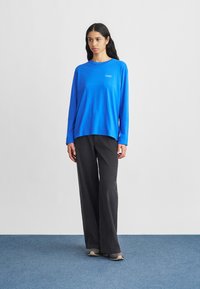 Μπλε βαμβακερό πουκάμισο με μακριά μανίκια και λαιμόκοψη στρόγγυλη, με λογότυπο, συνδυασμένο με σκούρα παντελόνια φαρδιάς γραμμής και γκρι αθλητικά παπούτσια.