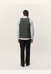 Grønstribet vest med en struktureret stof, båret over en lyseblå skjorte, parret med sorte bukser. Set fra ryggen.