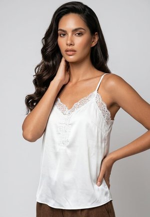 Mujer con cabello largo y oscuro, vistiendo un camisón de satén blanco con ribete de encaje, posando con una mano en el cuello y la otra en la cadera.