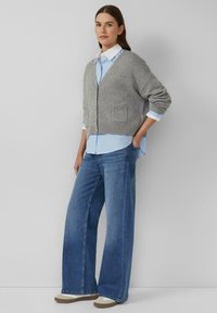 Frau, die einen grauen Knopf-Cardigan über einem blau-weiß gestreiften Hemd, weiten blauen Jeans und beigen Sneakers trägt und vor einer schlichten Wand steht.