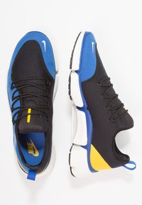 Nike tenisky s černým síťovaným svrškem, modrými a žlutými akcenty a bílou polstrovanou podrážkou. Obsahují šněrovací design a poutko pro snadné nazouvání.