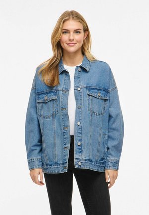 Młoda kobieta o długich blond włosach, ubraną w jasnoniebieską kurtkę dżinsową na białej bluzce i czarnych spodniach, stojąca na jednolitym tle.