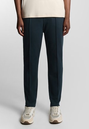 Marineblauwe op maat gemaakte broek met plooien; glad materiaal, elastische tailleband en manchetten; gecombineerd met sneakers in neutrale kleuren.