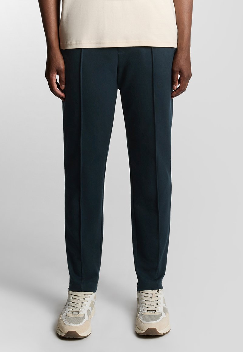 Marineblauen maßgeschneiderte Hosen mit Falten; glattes Material, elastischer Bund und Bündchen; kombiniert mit Sneakers in neutralen Farben.