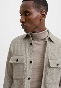 Lys beige tekstureret skjorte med to brystlommer, knapmansjetter og mørke knapper, lagdelt over en lys tan rullekravesweater.