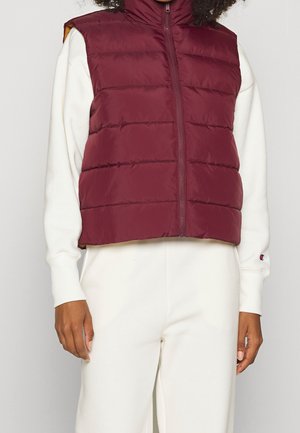 Persoon die een mouwloos bordeaux rood gewatteerd vest draagt over een wit sweatshirt met lange mouwen en een witte broek, staand tegen een effen achtergrond.