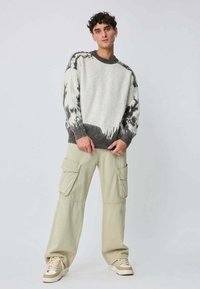 Jeune homme portant un pull à motifs tie-dye gris et blanc, un pantalon cargo beige et des baskets, se tenant contre un fond clair uni.