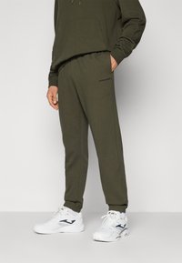 Ελαιόχρωμο joggers κατασκευασμένα από μαλακό ύφασμα, με ελαστική μέση και διακριτική επωνυμία στο πλάι. Συνδυασμένα με λευκά αθλητικά παπούτσια.