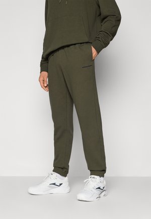 Olivgrüne Jogginghosen aus weichem Stoff, mit einem elastischen Bund und dezentem Branding an der Seite. Kombiniert mit weißen Sportschuhen.