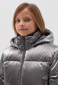 Silberne gepolsterte Jacke mit hohem Kragen, Frontreißverschluss und einer glänzenden, glatten Textur. Sie verfügt über eine Kapuze und ein gestepptes Pufferdesign.