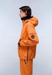 Ung person i profil iført en orange hættetrøje og matchende bukser med hænderne i lommerne mod en ensfarvet baggrund.