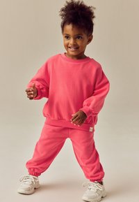 Roosa puuvillane pusa soonikkoes varrukate ja servaga, sobitatud samas toonis jogger-pükstega. Valged tossud, mänguline poos neutraalse tausta ees.