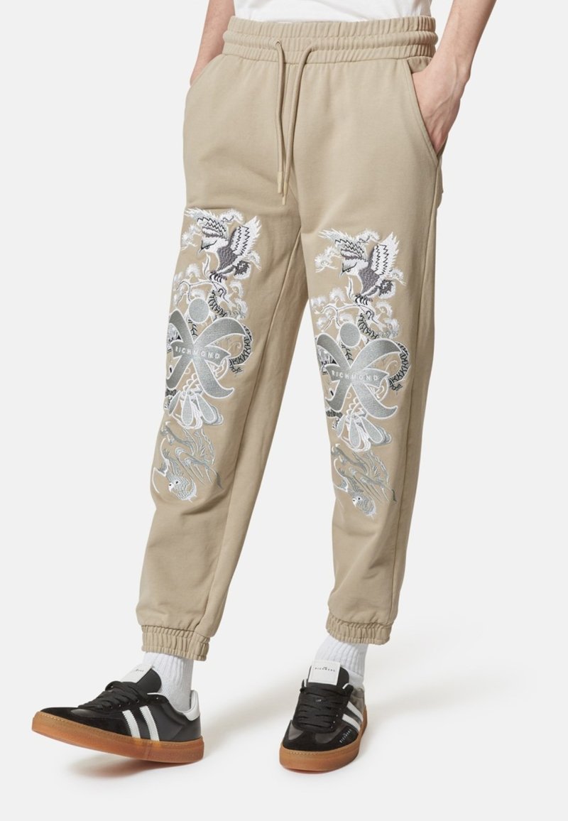 Pantaloni sportivi beige con vita e polsini elasticizzati, caratterizzati da un intricato ricamo argento e nero di uccelli e motivi su entrambe le gambe, abbinati a sneaker nere.