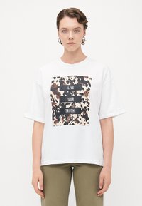 Person med kort hår som bär silverfärgade hoopörhängen och en vit t-shirt med ett brun-svart prickigt mönster samt texten "LEVA DIN SANNING".