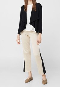 Cardigan noir ouvert sur un chemisier blanc, associé à un pantalon beige clair avec des bandes noires sur les côtés. Porté avec des chaussures plates marron clair.