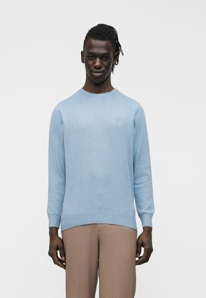 Homme debout portant un pull à col rond bleu clair et un pantalon marron clair sur un fond blanc uni.