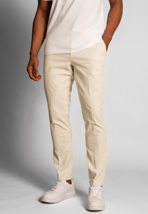 Pantalons beige fuselés en tissu léger, avec un devant plat, des poches latérales, associés à des baskets blanches.
