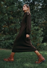 Brun strikket kjole med lange ærmer, ribstrikkede detaljer og gyldne accenter. Sammen med brune snørestøvler og tan-farvede sokker, placeret foran grønt løv.