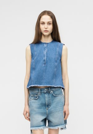 Noorpruuni juustega noor naine, kes kannab sinist varrukateta teksatoppi lukuga ja kõrge vöökohaga narmendavate teksapükstega valge tausta ees.