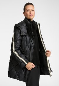 Mujer con chaqueta de abrigo acolchada negra con rayas blancas en las mangas, sosteniéndola abierta para mostrar el forro interior, vestida de negro debajo.