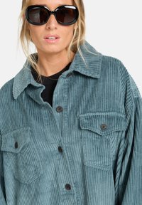 Teal corduroyjas met opvallende verticale ribbels, twee borstzakken en zwarte knopen. Model draagt een oversized zwart zonnebrillen.