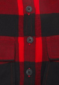 Camicia a scacchi rossa e nera con apertura a bottoni, due tasche sul petto e cuciture visibili. Il tessuto sembra morbido e texturizzato.