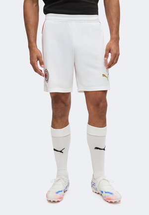 Mand iført hvide AC Milan fodboldshorts med gyldent Puma-logo, hvide Puma-strømper og hvide fodboldstøvler med røde og blå accenter.