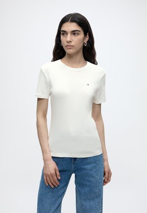 Γυναίκα με μακριά σκούρα μαλλιά, που φορά μικρούς κρίκους σκουλαρίκια, εφαρμοστή λευκή κοντομάνικη μπλούζα με μικρό λογότυπο και μπλε τζιν, σε λευκό φόντο.