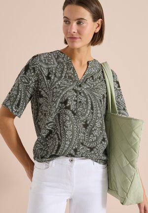 Femme portant une chemise verte à motif cachemire et un pantalon blanc, tenant un sac cabas matelassé vert clair sur son épaule.
