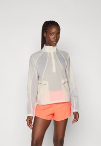 Veste coupe-vent légère et translucide avec un col haut, deux poches avant et des poignets ajustables ; associée à un short corail vif.